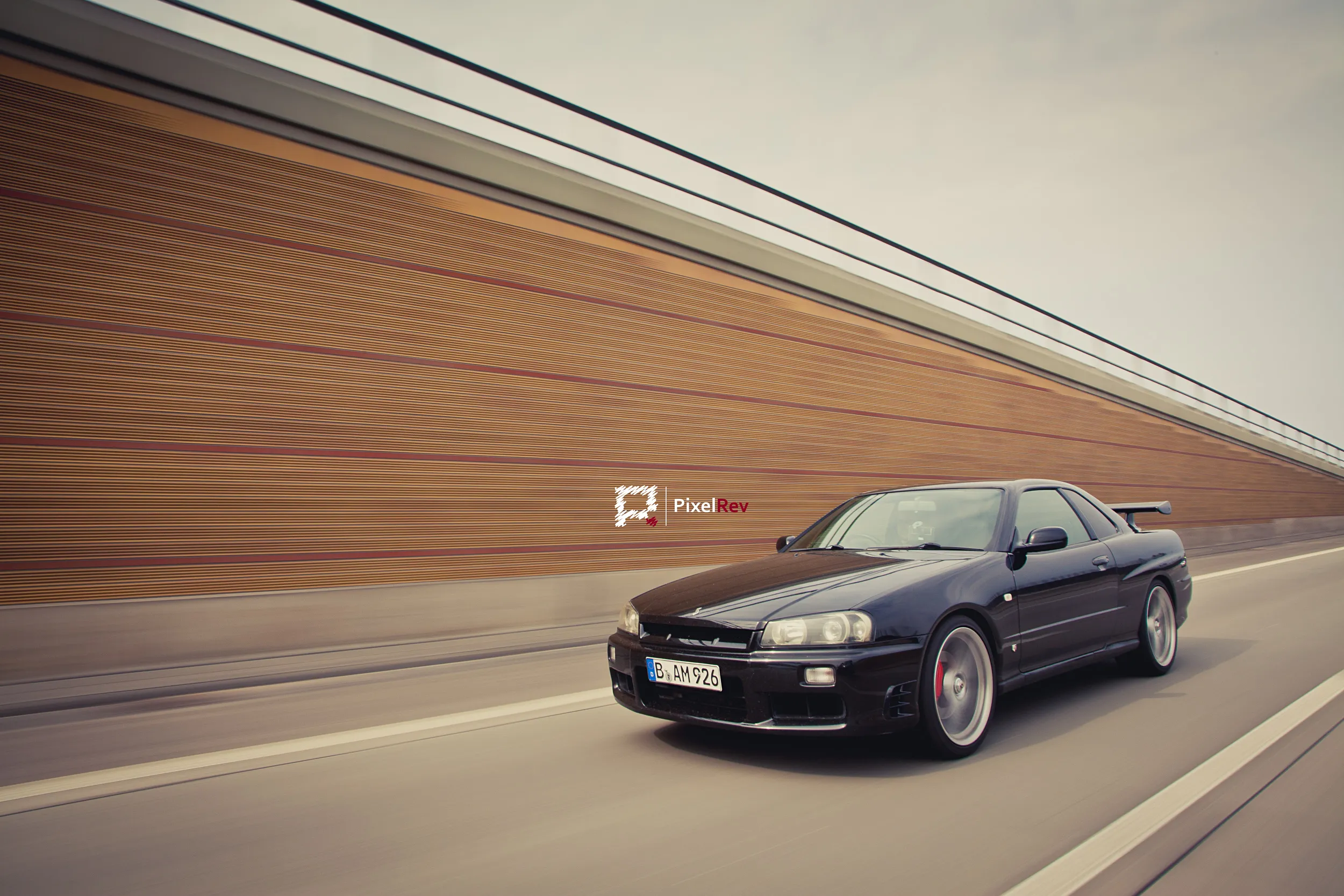 Dunkler Nissan Skyline R34 GT-R in Fahrt vor einer verwischten Lärmschutzwand