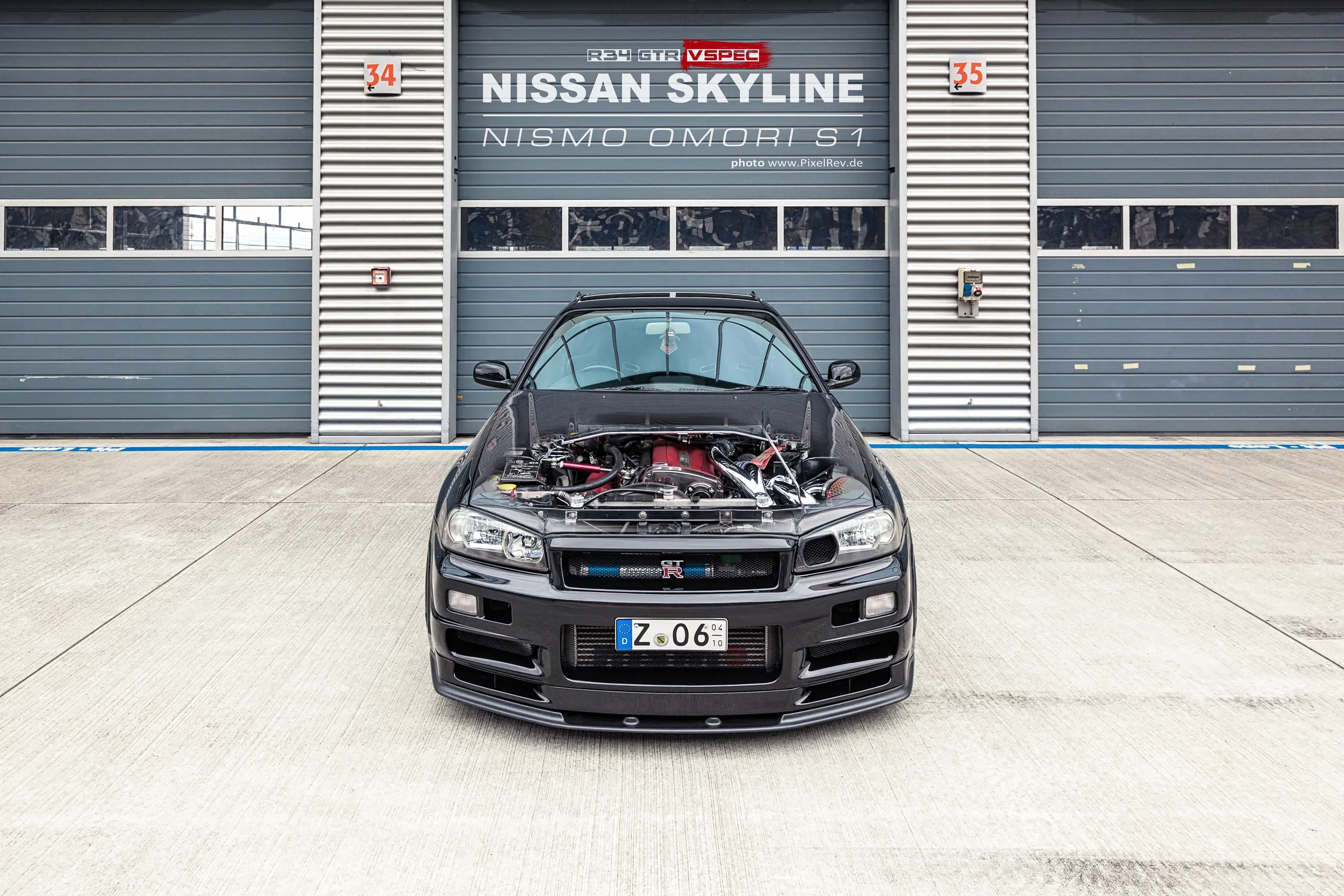 Nissan Skyline R34 GT-R Nismo Omori S1 mit geöffneter Motorhaube vor einem Rolltor mit Titel