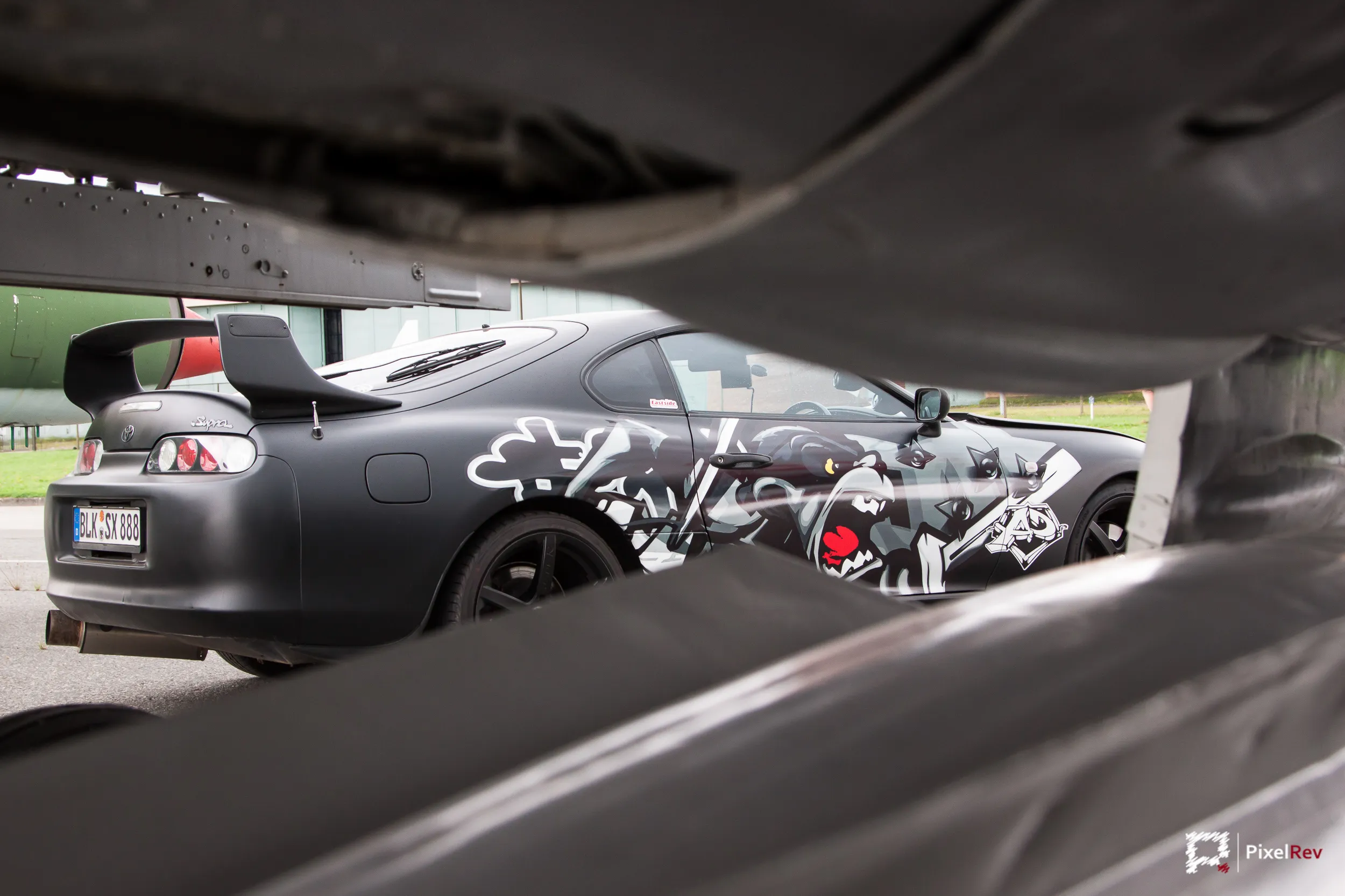 Folierter Toyota Supra, unter dem Rumpf eines Kampfflugzeugs hindurch fotografiert
