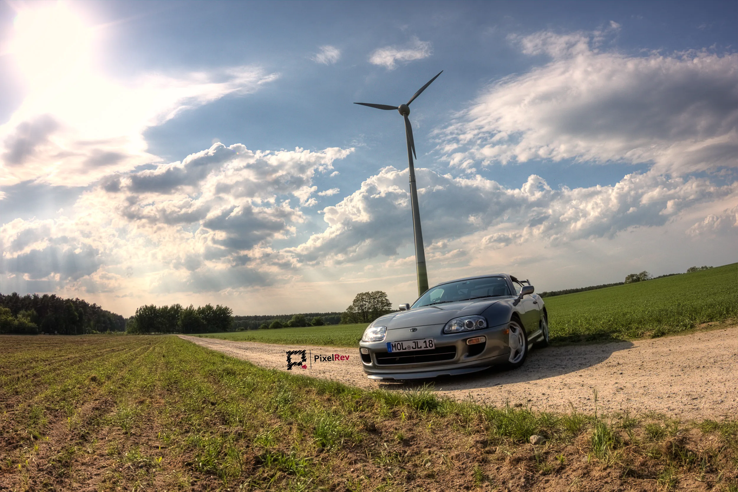 Silberner Toyota Supra Mk4 auf einem Feldweg unter dramatischem Wolkenhimmel mit Windrad