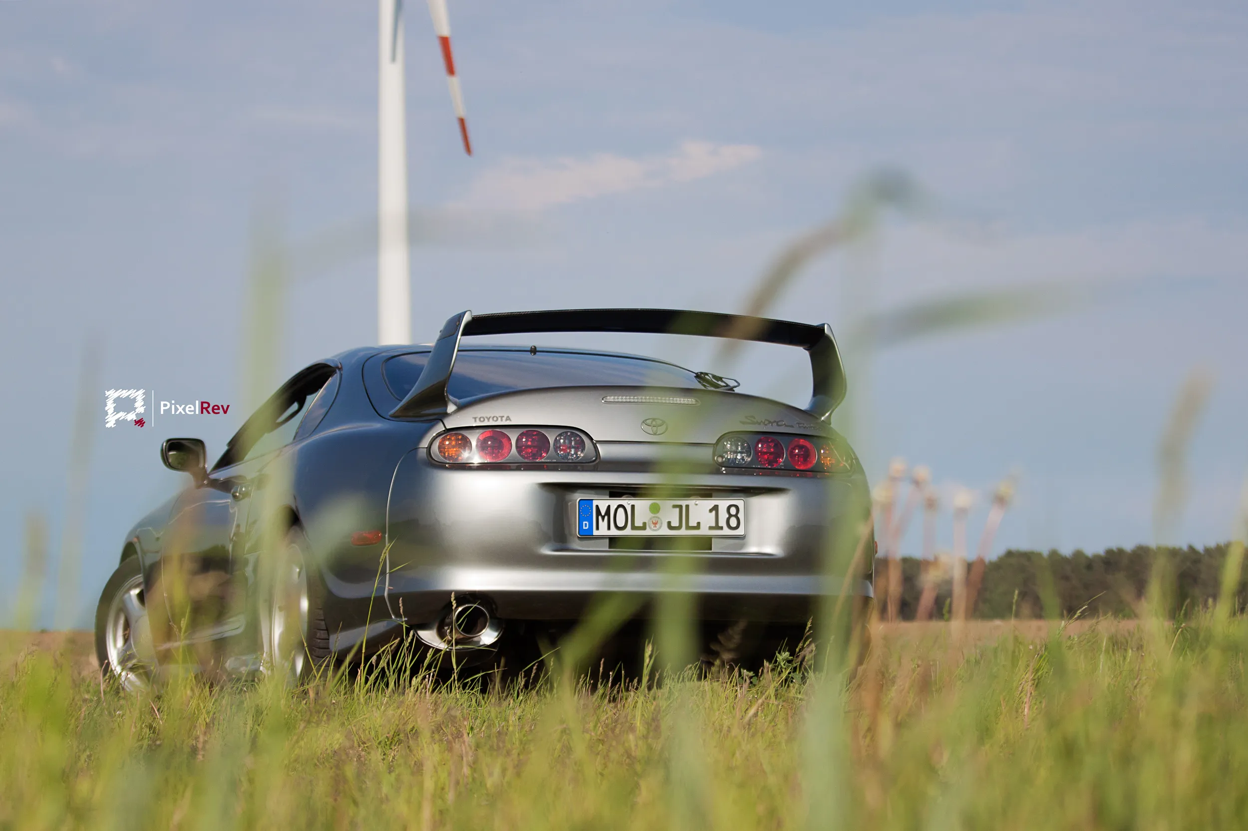 Heck eines silbernen Toyota Supra Mk4 mit Heckflügel, durch hohes Gras fotografiert