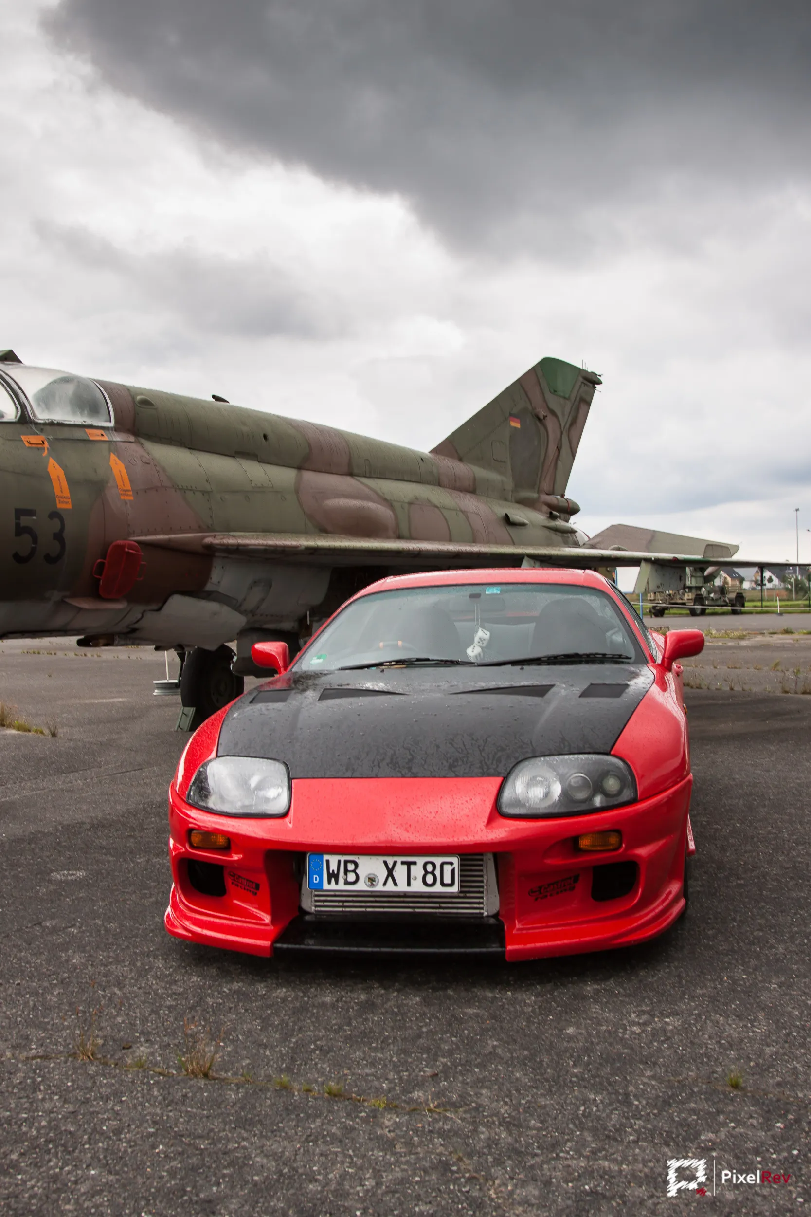 Roter Toyota Supra mit Carbon-Motorhaube frontal vor einem Kampfjet